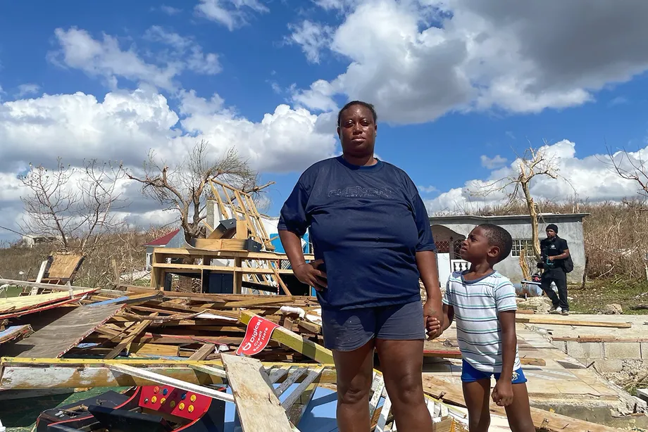 Una mujer y un niño se encuentran entre maderas rotas y escombros tras los daños causados por la tormenta. Bajo un cielo despejado pero cubierto de nubes, sus rostros reflejan la dureza de lo vivido y la fortaleza para seguir adelante.