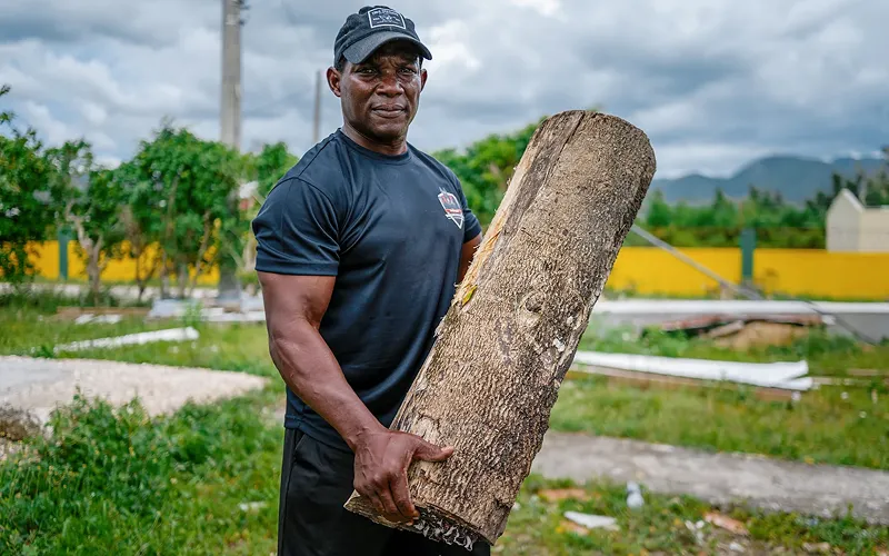 Un hombre sonriente, vestido con ropa de trabajo oscura y una gorra, sostiene un grueso tronco de árbol en un entorno al aire libre. Al fondo se distinguen vegetación, un muro amarillo y montañas.