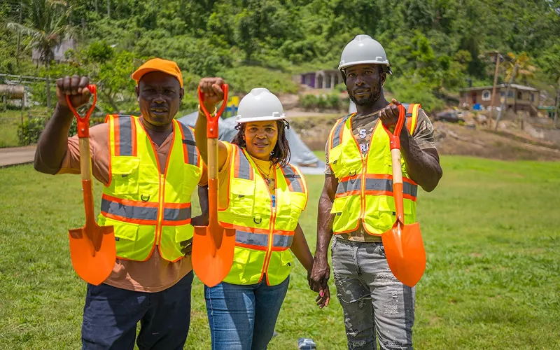 Tres trabajadores de la construcción posan en una zona rural con cascos y chalecos reflectantes, sosteniendo palas de color naranja. Sus expresiones y actitud reflejan orgullo por su trabajo y un claro espíritu de equipo.