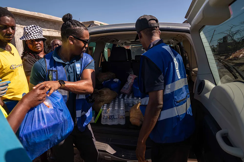 Personal humanitario con chalecos azules descarga botellas de agua y otros artículos de primera necesidad de un todoterreno y entrega una bolsa azul a varias personas en un lugar concurrido al aire libre en un día soleado.