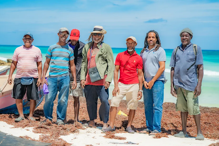 Un grupo de adultos posan juntos en una playa soleada, con el mar turquesa de fondo. Su actitud relajada transmite un ambiente de confianza y camaradería.