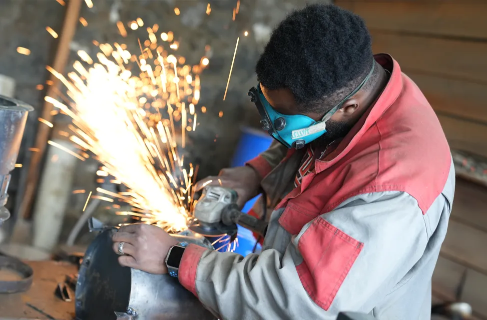 Sparks are seen flying out as a man wearing blue safety goggles works on a metal project in a workshop setting.