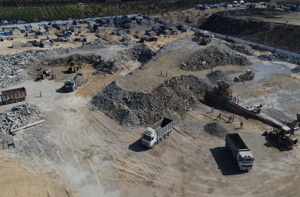 Aerial view of a construction site with piles of rubble and gravel. Trucks and machinery are moving materials. Workers in orange vests are visible.