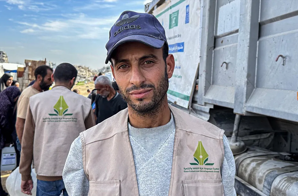 Man wearing a blue cap and beige vest with a logo stands in front of a truck. He appears focused. Others in similar vests are in the background.