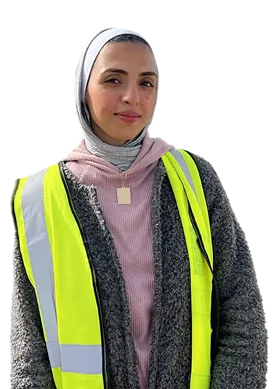 A woman in a yellow safety vest and gray coat. She wears a light hijab and a calm expression, exuding confidence.