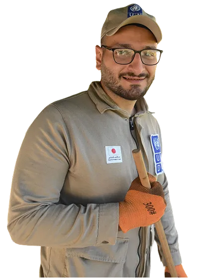 A man in a beige uniform and cap smiles, holding a shovel. He wears glasses and orange gloves, with UNDP and Japanese flag logos on his jacket.