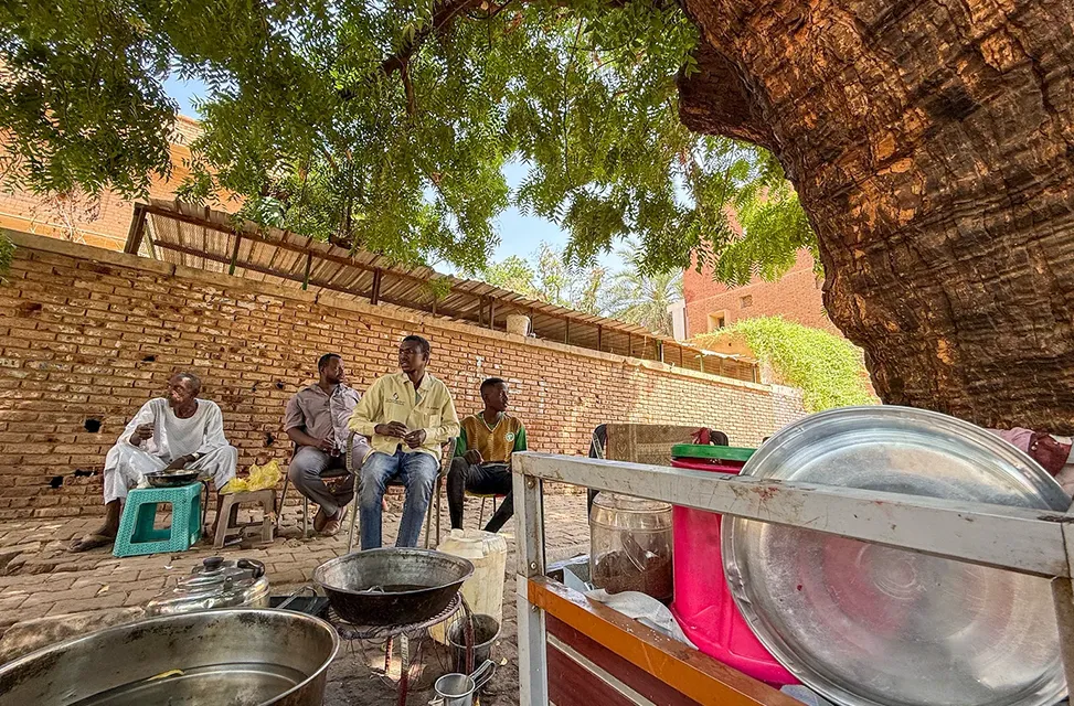 Four men sit and talk under a large tree next to a brick wall; cooking pots, utensils and a food cart are in the foreground.