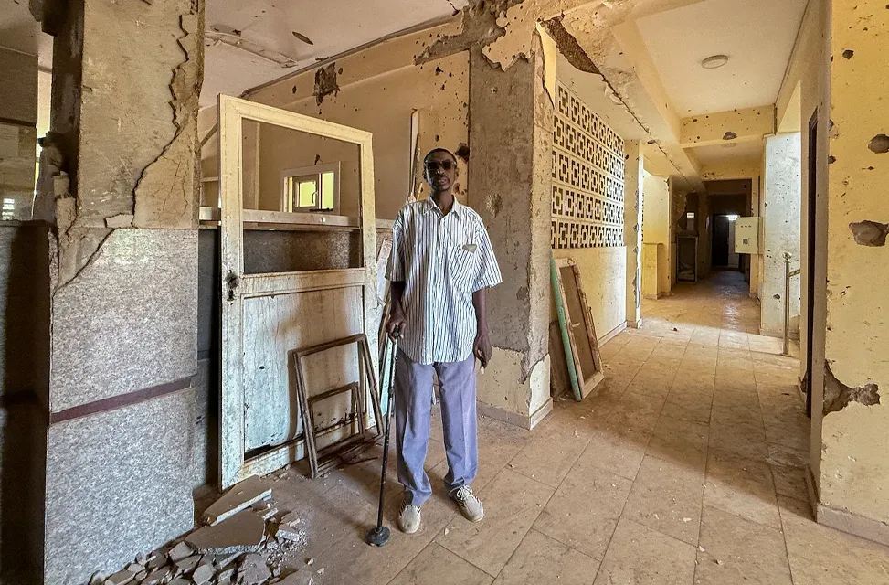 A man stands in a large room with damaged stone and tile walls and debris on the floor.