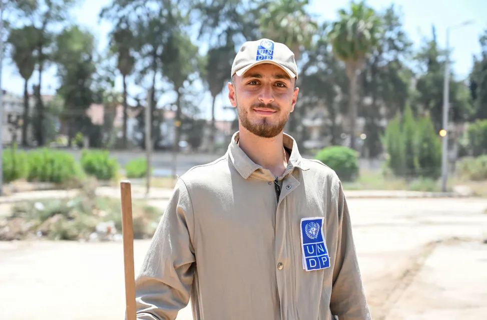 A smiling man in a beige uniform and cap with UNDP logos stands holding a long wooden handle in one hand, with trees and a blurred landscape in the background.
