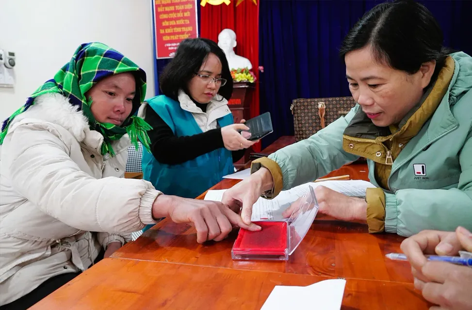 A woman in a green headscarf presses her finger into a red ink pad at a wooden table, assisted by another woman.
