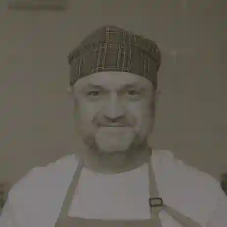 No Color-Middle-aged man wearing a checkered chef’s hat and a yellow apron, standing in a kitchen.