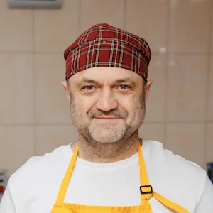 Hombre de mediana edad con un gorro de chef a cuadros y un delantal amarillo, de pie en una cocina.