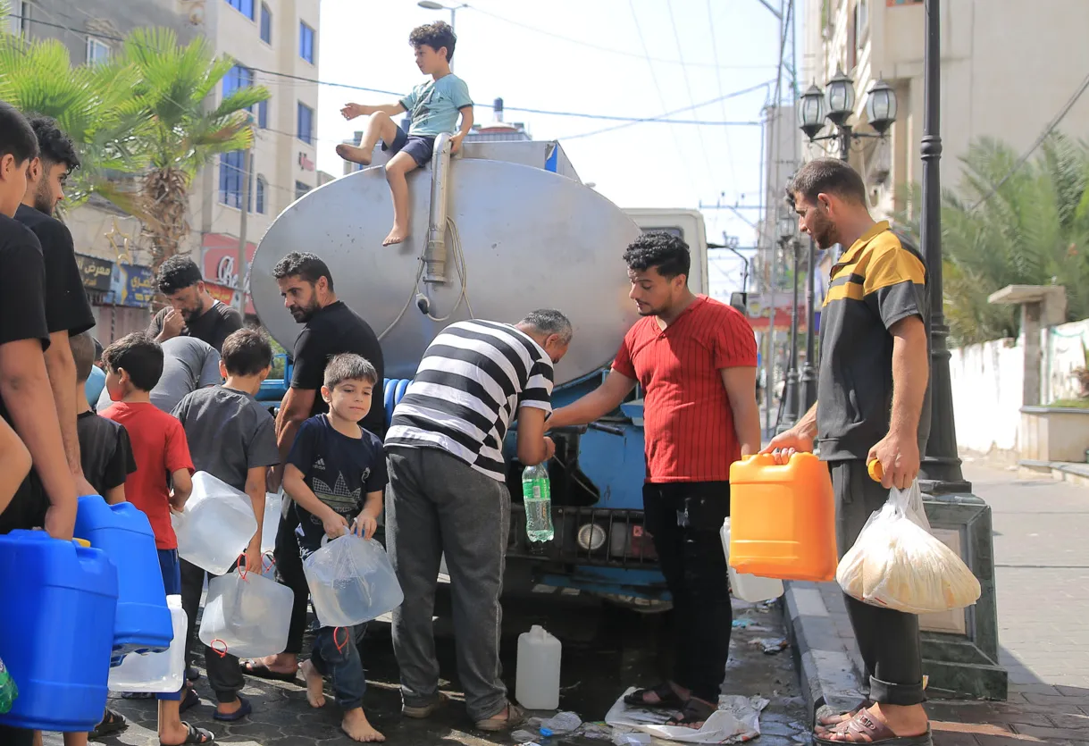 Un grupo de niños y adultos se reúnen alrededor de un camión de suministro de agua en una calle, mientras llenan recipientes en un entorno urbano soleado.