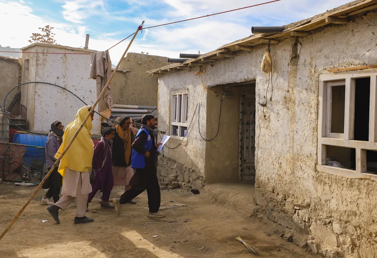 A group of people walk along a dirt pathway near weathered, mud-walled buildings with open windows.