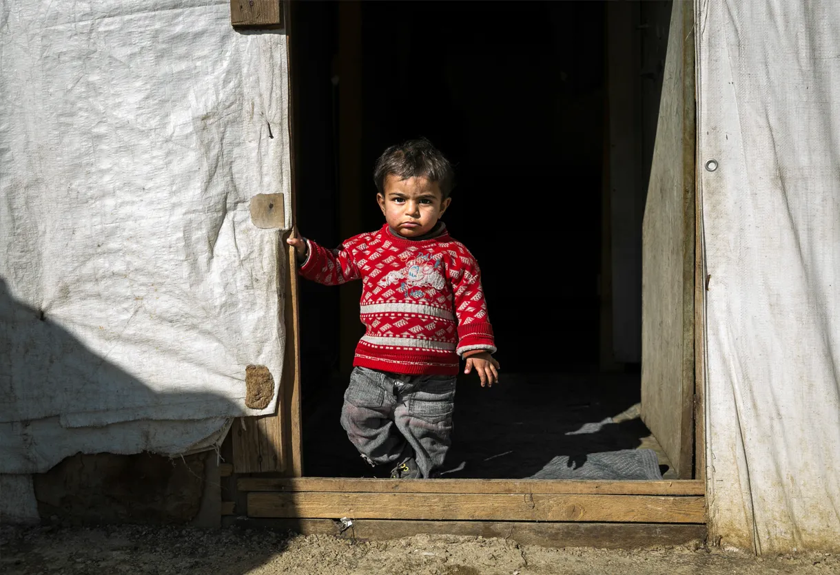 A young child in a red sweater stands at the entrance of a makeshift shelter, with sunlight illuminating the scene.