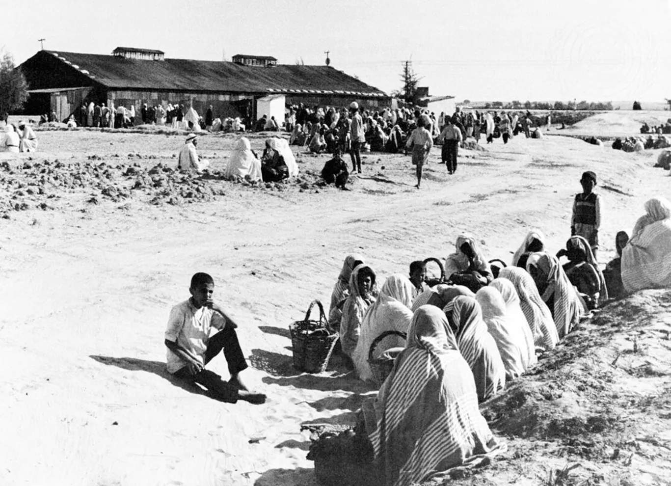 A group of seated individuals in traditional attire gather in a dusty area, with a building and more people in the background.