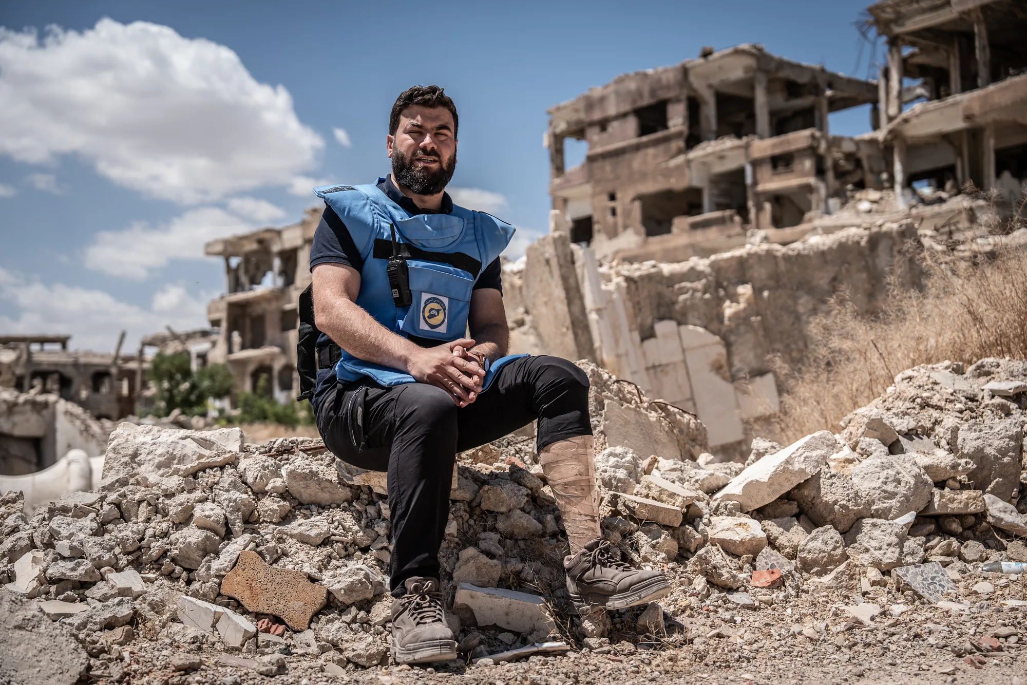 Bearded man in a blue protective vest sits amid rubble, with war-damaged buildings behind him under a bright sky.