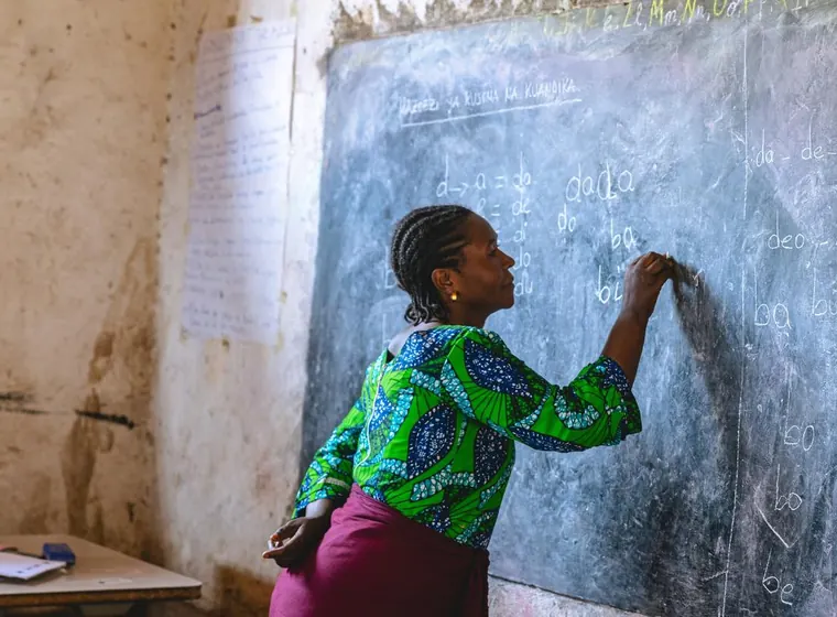 A teacher writes on a chalkboard at the front of a classroom.