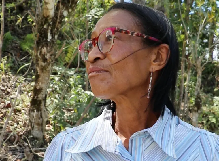 A woman wearing glasses and earrings looks into the distance, with trees behind her.
