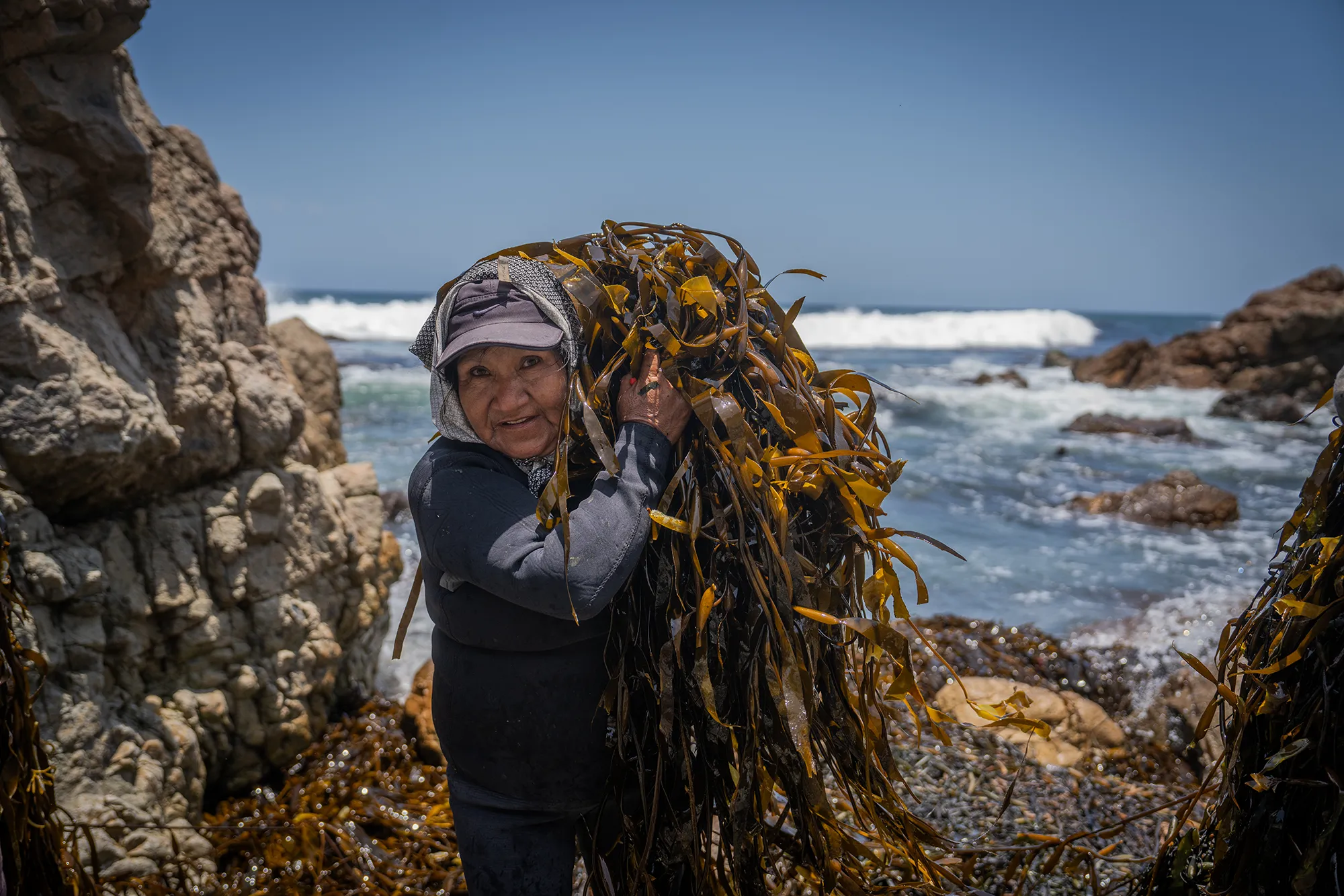 Una mujer se encuentra en una orilla rocosa con un gran bulto de algas al hombro. Lleva sombrero y ropa protectora, sonriendo levemente mientras las olas rompen al fondo.