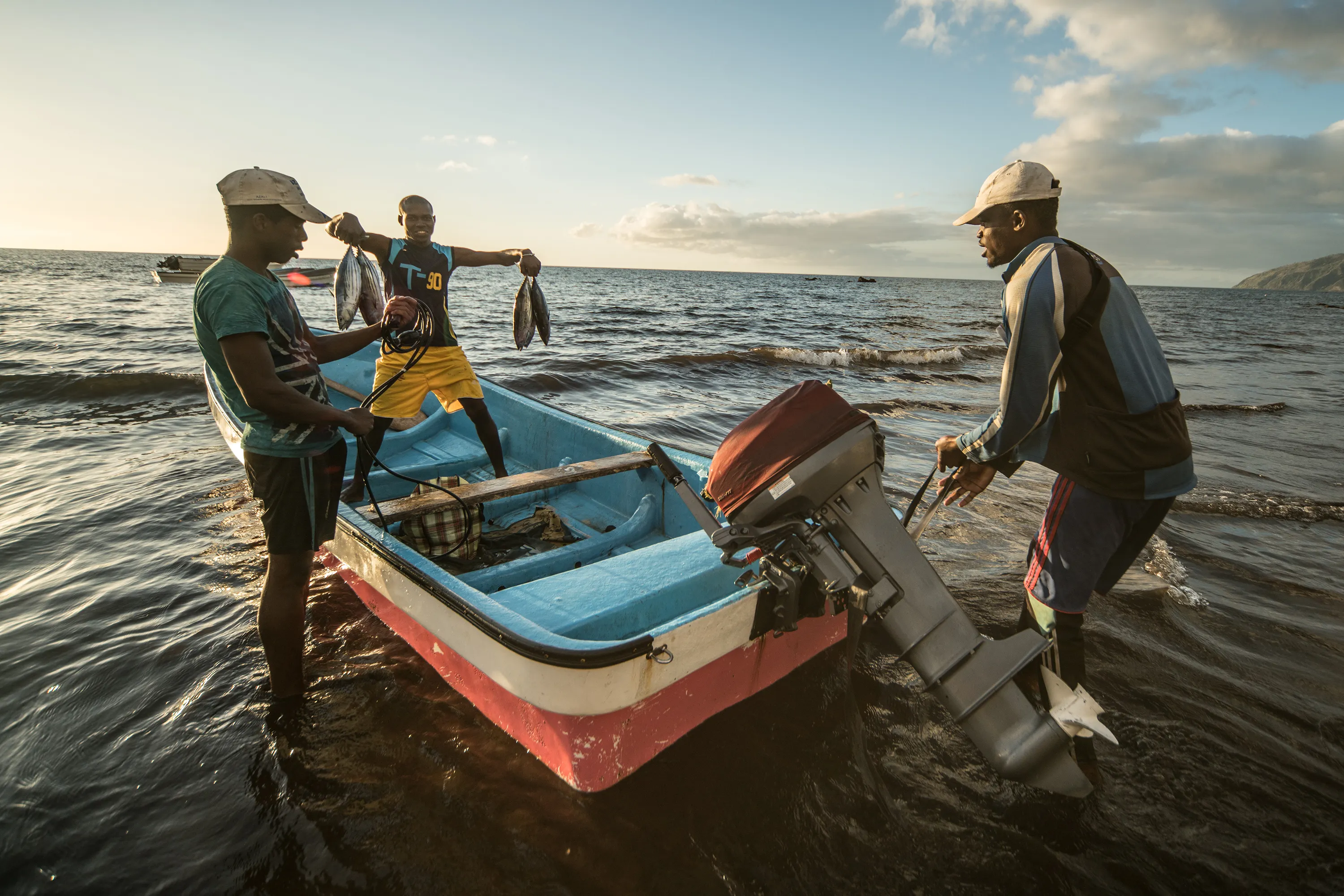 Tres pescadores en las Comoras se encuentran junto a una pequeña lancha a motor en la orilla al atardecer. Un hombre dentro de la lancha sostiene con orgullo el pescado recién capturado, mientras los demás ayudan con el equipo y el motor fueraborda. El océano se extiende tras ellos, reflejando la luz dorada.