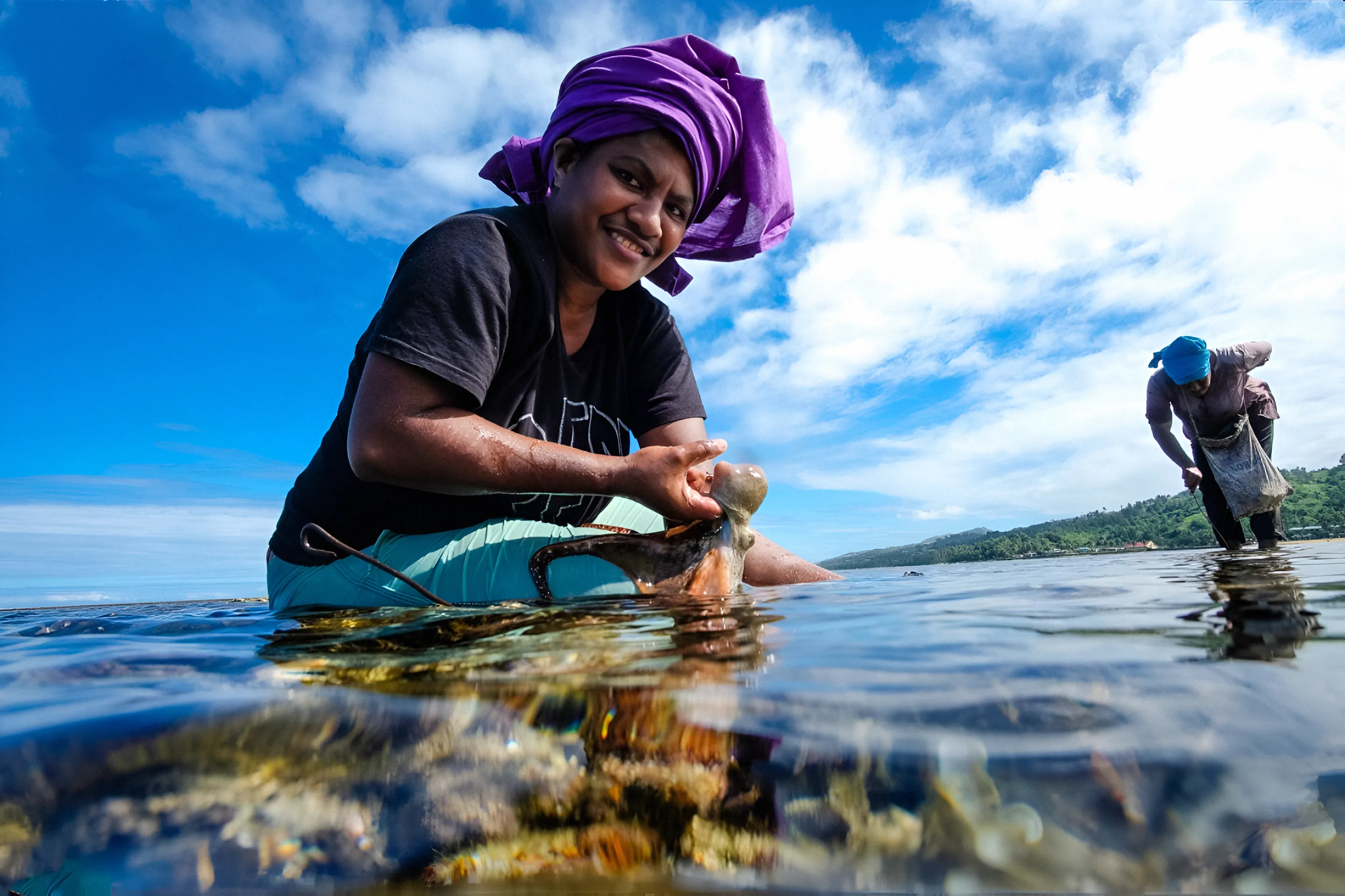 Una mujer con un pañuelo morado sonríe mientras se arrodilla en aguas costeras poco profundas, sosteniendo un pulpo recién capturado. Al fondo se ve a otra mujer, también vadeando y pescando. El agua cristalina revela corales, con una exuberante costa verde y un cielo azul a lo lejos.