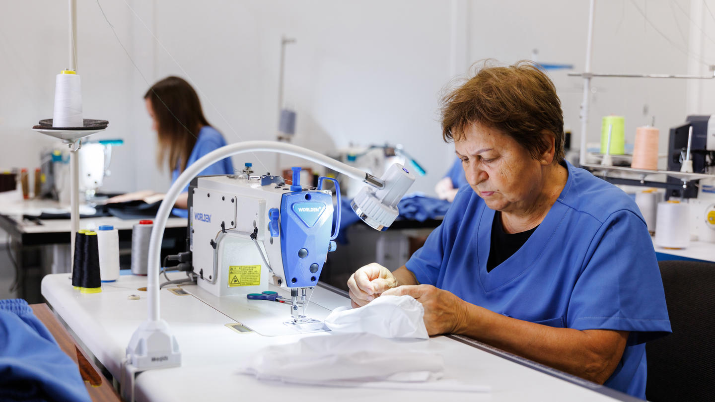 Focused sewer in a blue shirt operates an industrial sewing machine, while a coworker works in the bright textile workshop behind her.