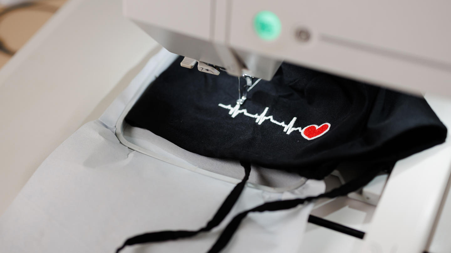 Embroidery machine stitching a white heartbeat line and red heart onto black fabric.