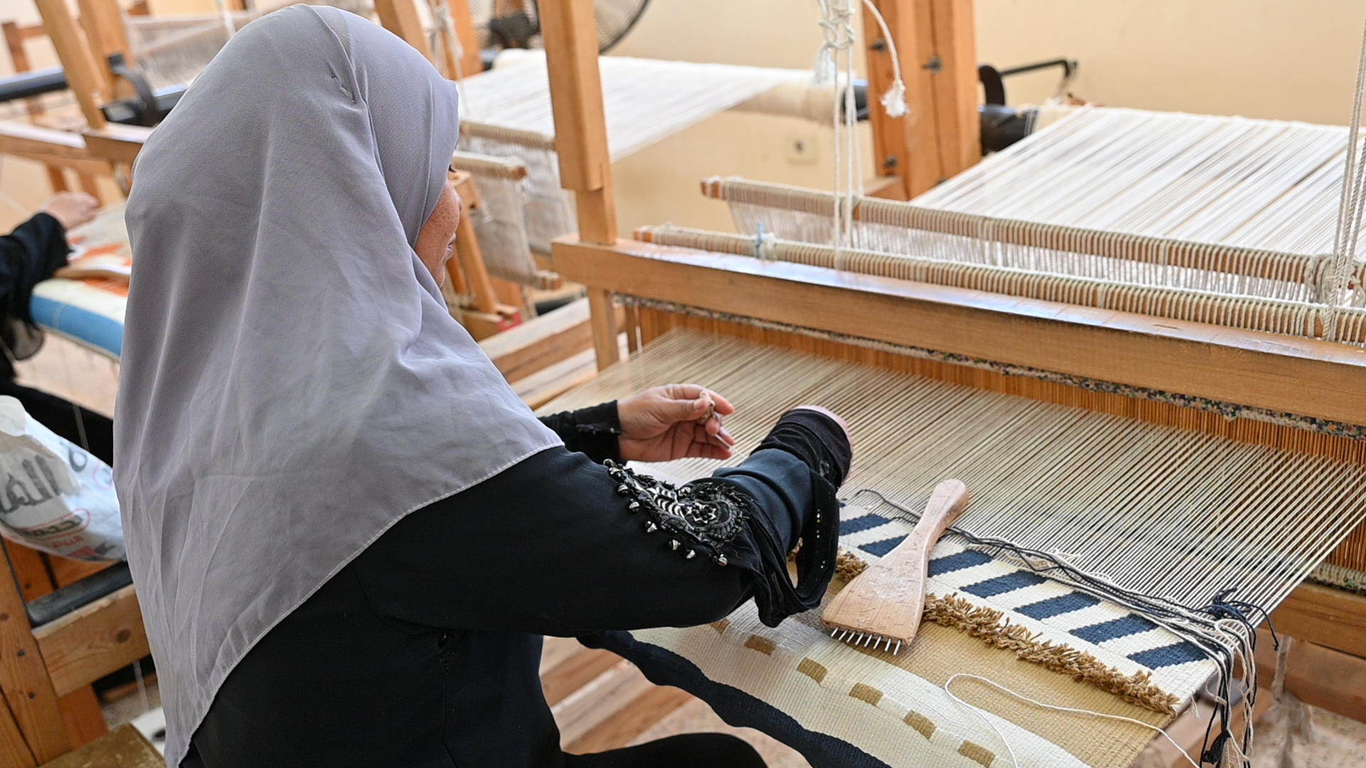 Woman in a gray hijab weaves a patterned textile on a wooden loom.