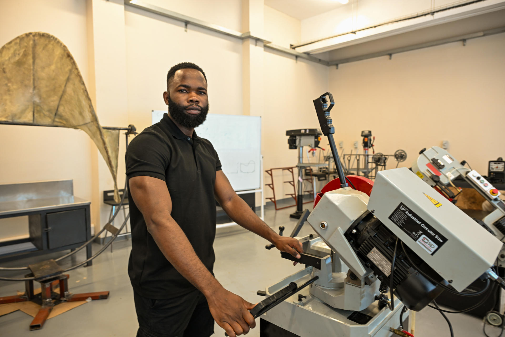 A bearded man in a black polo operates a large industrial machine a bright workshop.