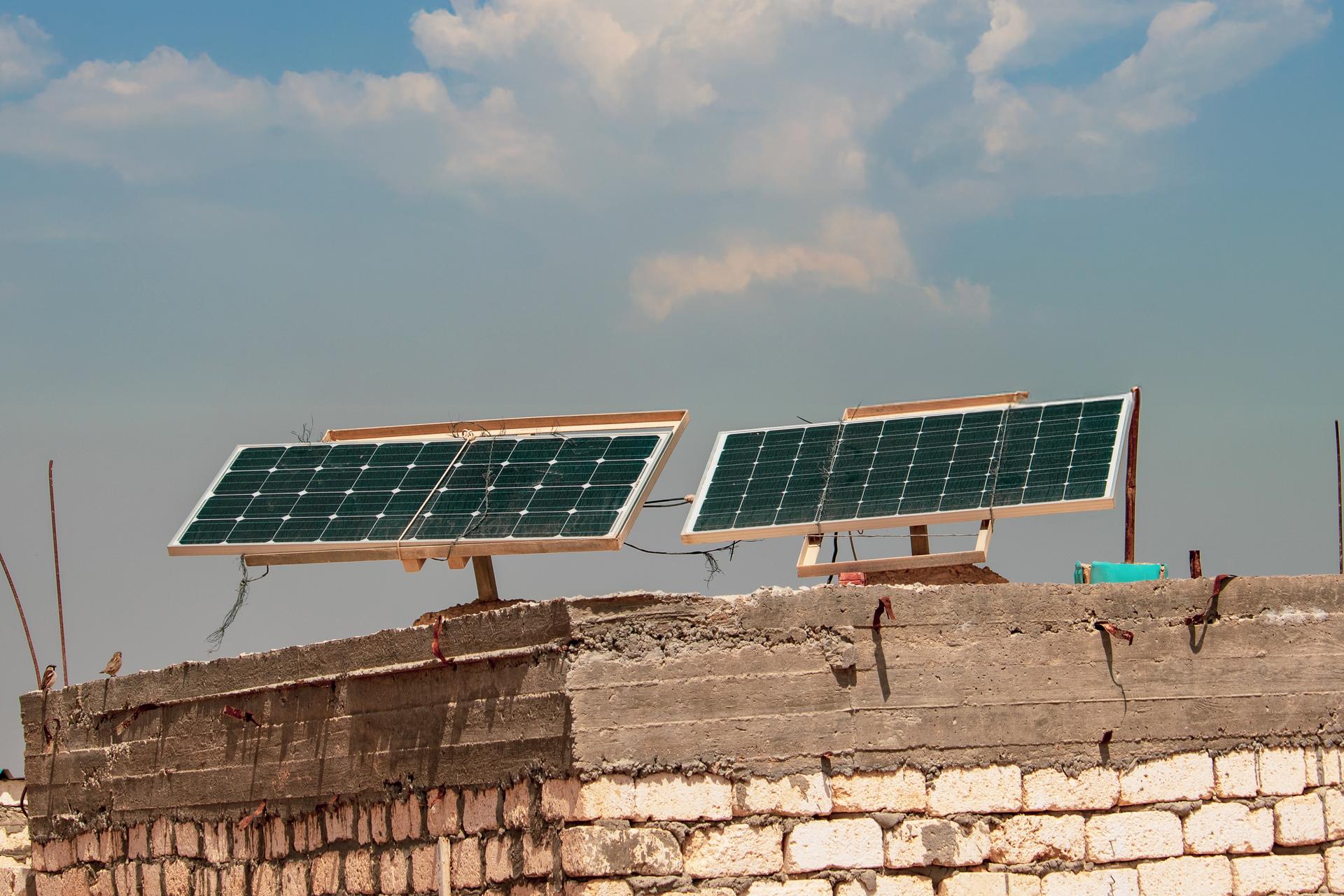 Two solar panel arrays sit on a rustic stone wall under a bright blue, cloud-dotted sky.