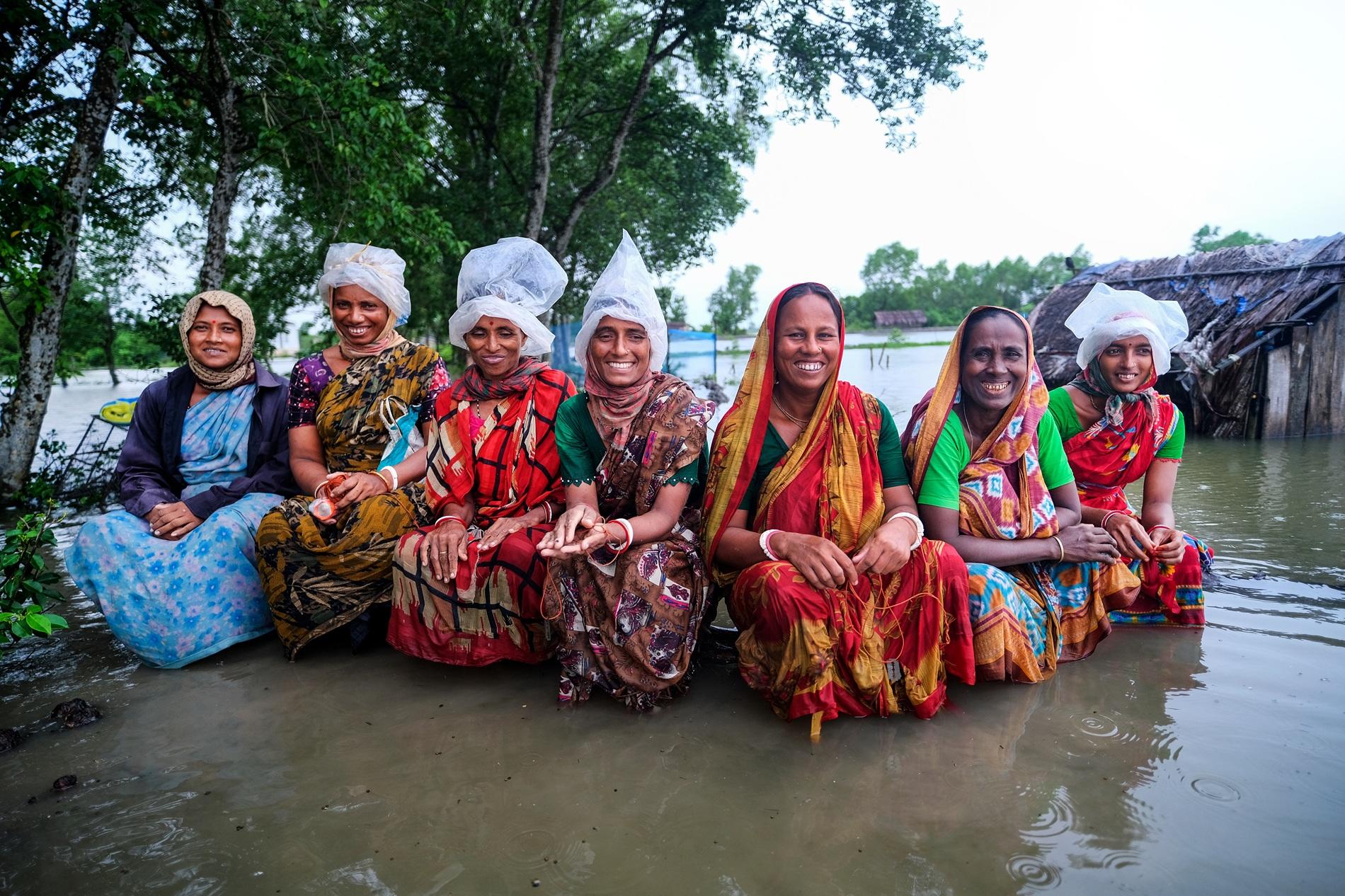 Seven women in colourful saris and headscarves sit with their feet in water beside riverside homes.