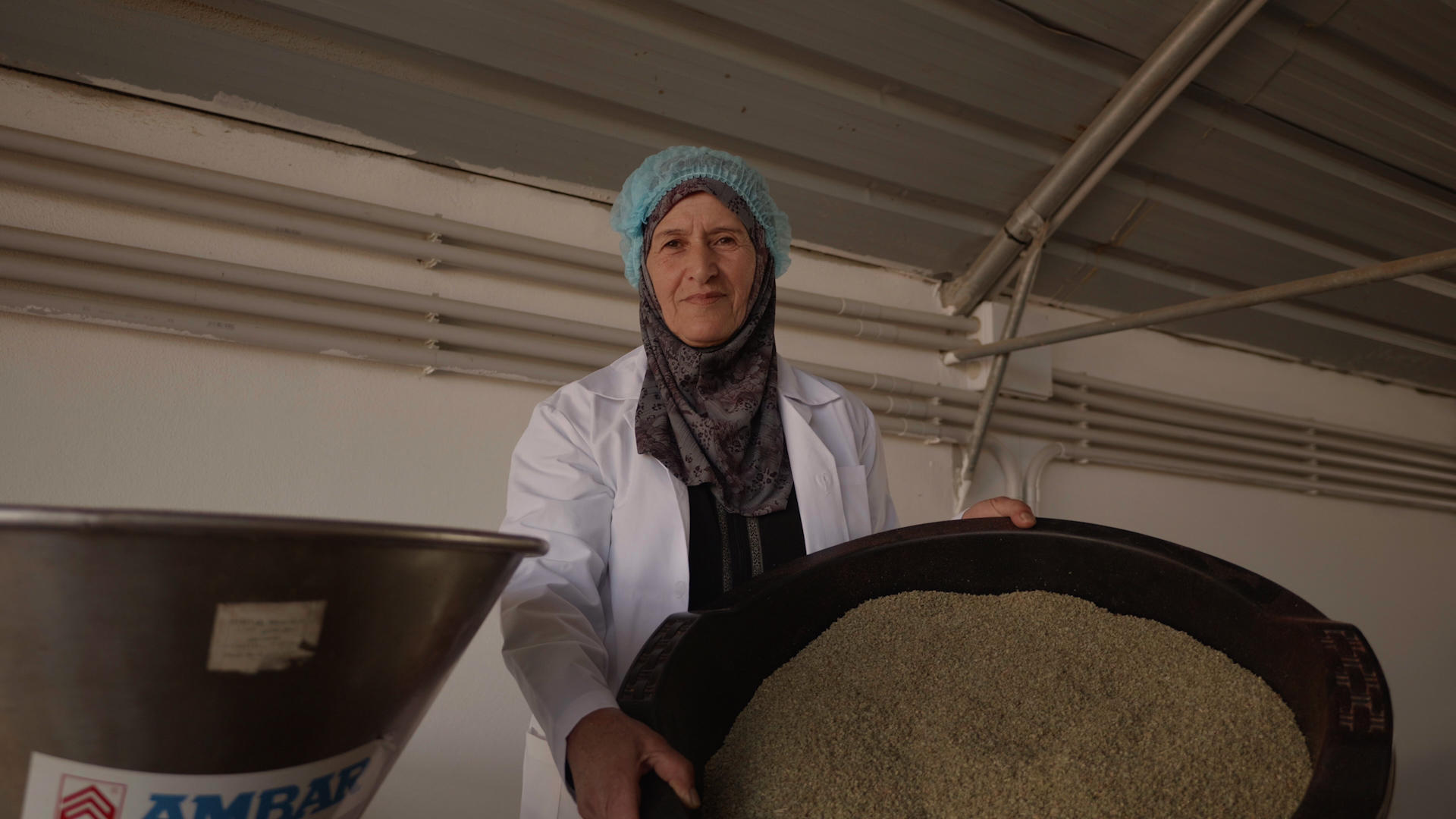 Woman in a white lab coat and headscarf holds trays of grain in an industrial facility.